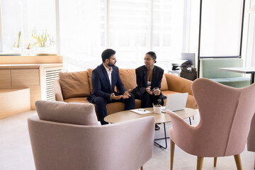 Young multiethnic couple of Indian and African businesspeople talking on sofa in co-working space cozy interior, discussing collaboration, enjoying offline communication, networking
