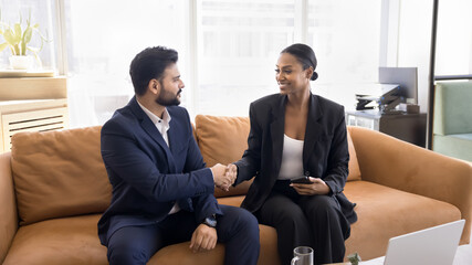 Two happy diverse businesspeople shaking hands at co-working workplace with couch, smiling, enjoying teamwork, communication, professional friendship, sitting on sofa in office hall