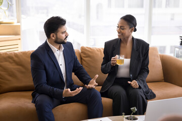 Two multiethnic business man and woman discussing work tasks, plan, project strategy over cup of tea, sitting on comfortable couch, in co-working room, talking, smiling