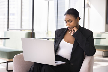 Thoughtful beautiful African business specialist woman using laptop for communication, sitting in armchair in co-working office, touching chin, reading text on screen, watching content, thinking