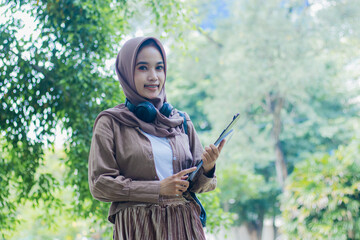 Young indonesia woman in veil candid smile carrying book and clipboard standing in outdoor park. female students for the themes of education, technology and advertising