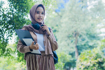 Young asian girl in hijab smiling carrying book and clipboard standing holding hijab in outdoor...