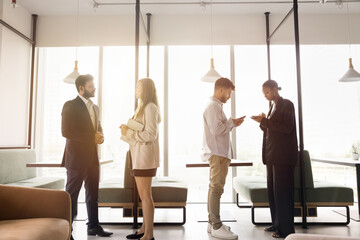 Two multiethnic couples of office colleagues in formal clothes discussing work projects, standing and talking in co-working office space, speaking, using gadgets. Full length side view shots