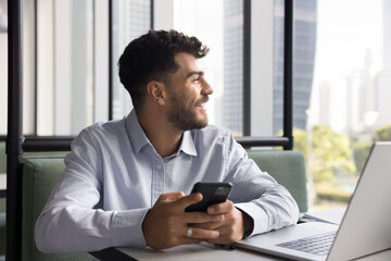 Cheerful young Arab business project manager man using Internet technology for work communication, typing on mobile phone at laptop, holding smartphone, looking at window away, smiling