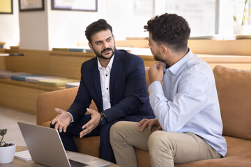 Confident young Indian business man presenting project on laptop to client, customer, partner, pointing at display, talking to colleague on office couch. Professional mentoring new employee