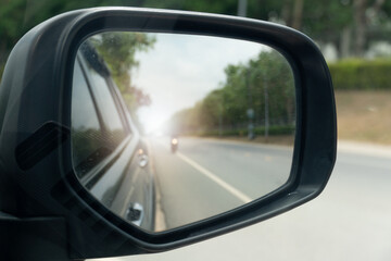 Inside view of mirrors wing. Rear view of a gray car with asphalt road and green trees in the daytime. Blurred image of a motorcycle approaching from behind.