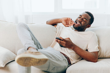 Happy African American man sitting on a black sofa, typing a message on his smartphone The modern...
