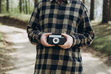 A man in a checkered shirt holds a film camera in his hands in the forest.
