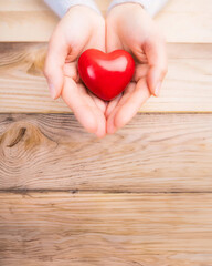 Cupped hands gently presenting a glossy red heart on a wooden background, symbolizing love and care.