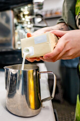 Barista skillfully pouring milk from a carton into a stainless steel pitcher, preparing for latte art in a cozy café setting
