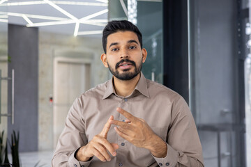A professional Hispanic businessman looking into the camera, engaging with viewers during a video call in a modern office environment.