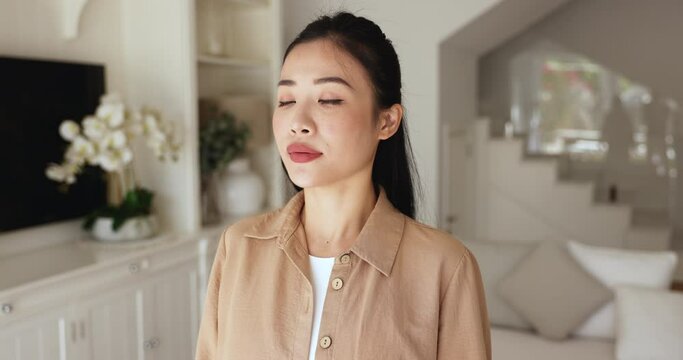 Close up Asian woman makes inhales exhales standing alone in living room with eyes closed, breathing fresh air inside, recover inner calmness after situation, reduces stress, take moment to revitalize