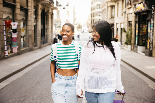 A young female couple is exploring a new city during a short weekend trip, walking together while pulling a suitcase along a city street.