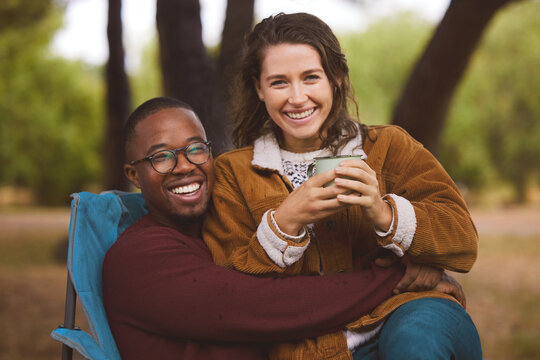 Couple, chair and love with camping in woods for weekend, fun adventure and interracial. Happy man, woman and sitting in forest for happiness with green trees, nature holiday and coffee with mug