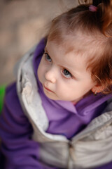 A pensive toddler looks up, her blue eyes thoughtful and emotive, set against the soft backdrop of her playful purple hoodie