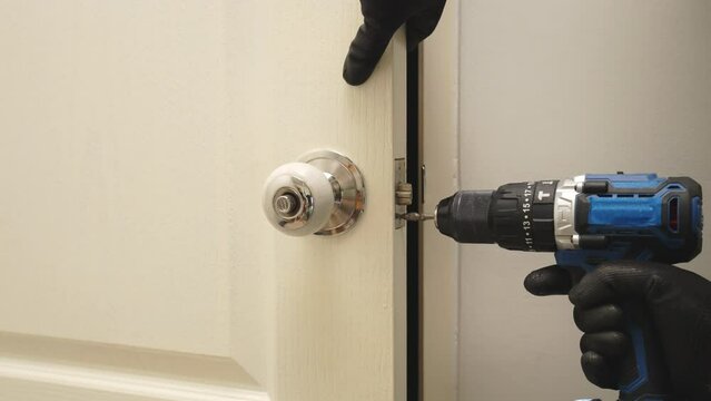 A man is using a power drill to open a door. The door is white and the man is wearing black gloves