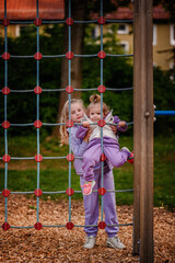 Two sisters share an adventurous moment on a climbing net at the playground, with a backdrop of woodchips and greenery enhancing their playful exploration