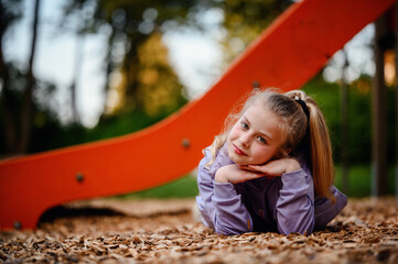 A young girl rests her head in her hands on the playground woodchips, her thoughtful gaze and the warm dusk light painting a picture of peaceful contemplation