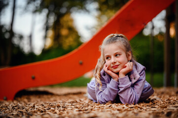 A young girl rests her head in her hands on the playground woodchips, her thoughtful gaze and the...