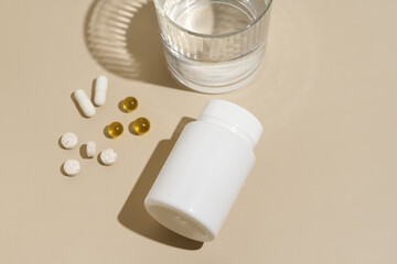 White mockup of blank jar and capsule pills on beige isolated background with glass of water. Health care, disease treatment, vitamins for beauty concept.