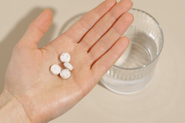 A woman's hand holds white pills before consumption against the background of a glass of water and a beige background. Concept of daily intake of medicines, vitamins, dietary supplements.