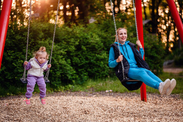A little girl and a woman enjoy swinging in a park at dusk, with golden sunlight filtering through trees in the background.