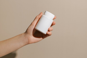 Female hand holding white small pill jar mockup on beige isolated background. Concept of pharmacy, health care, medicine, treatment. Image for your design.