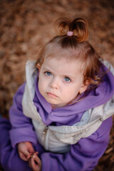 A close-up portrait of a thoughtful toddler in a purple jumpsuit, sitting on a playground, with...