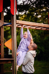 A girl reaches for the sky on a playground, supported by a smiling adult beneath her, capturing a...