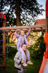 A joyful girl sits atop an adult's shoulders, reaching for the monkey bars at a playground, their smiles suggesting a shared moment of fun