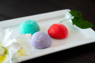 Three mochi different tastes and colors on white plate with flowers on black background. Japanese traditional frozen delicious dessert mochi. ice cream with dough of sticky rice. Asian cuisine.