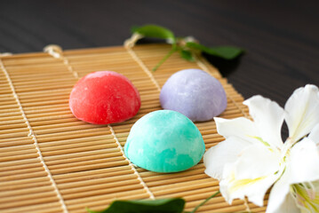 Three mochi different tastes and colors on bamboo makisu with flowers on black background. Japanese traditional frozen delicious dessert mochi. ice cream with dough of sticky rice. Asian cuisine.