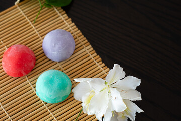 Fototapeta na wymiar Three mochi different tastes and colors on bamboo makisu with flowers on black background. Japanese traditional frozen delicious dessert mochi. ice cream with dough of sticky rice. Asian cuisine.