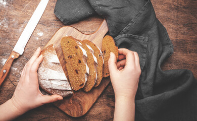 Women's hands holding rye sourdough bread with raisins, sliced pieces on wooden cutting board, bread knife and kitchen linen towel. Rustic table background, top view
