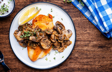 French breaded chicken steak with mushrooms and onion sauce with white wine, lemon and thyme on plate, rustic table background, top view