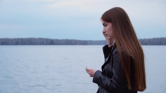 A young girl on the shore of the lake listens to a message and answers it on her mobile phone. The girl is chatting on a social network.