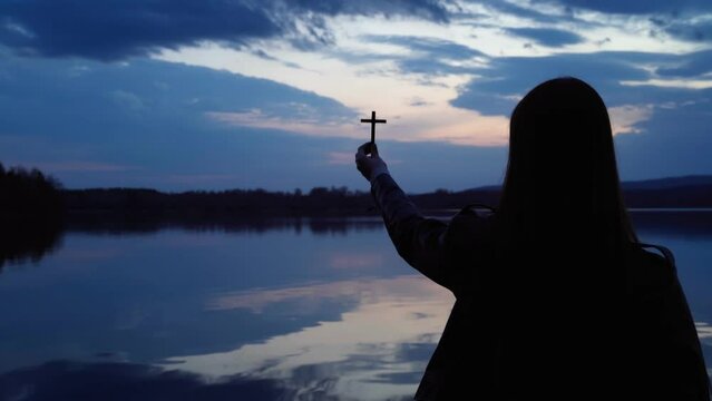 The silhouette of a girl raising an Orthodox cross to the sky at dusk. A mirror image of the cloudy sky on the surface of the water after sunset. The concept of religion and faith in God.