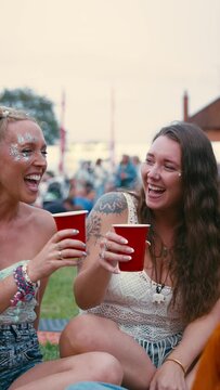 Vertical video of three female friends wearing glitter and doing cheers with drinks talking and having fun at outdoor summer music festival - shot in slow motion 