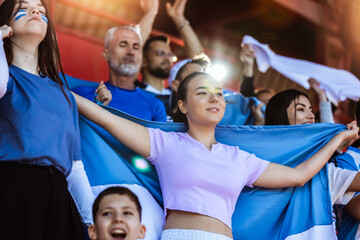 Sport fans cheering at the game on stadium. Wearing blue and white colors to support their team. Celebrating with flags and scarfs. - 790182485