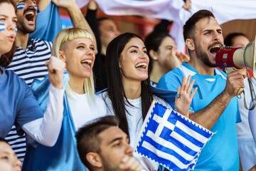 Greece fans cheering at the game on stadium. Wearing blue and white colors to support their team. Celebrating with flags and scarfs.