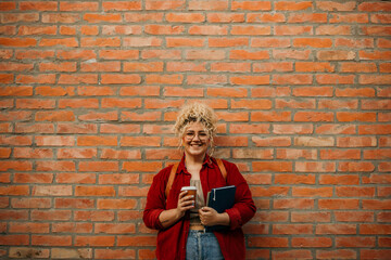 Portrait of a caucasian plus size woman with backpack holding coffee to go and books, standing on a...