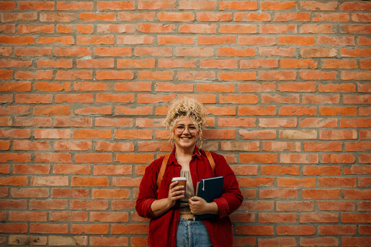 Portrait of a caucasian plus size woman with backpack holding coffee to go and books, standing on a city street. Caucasian student at the campus