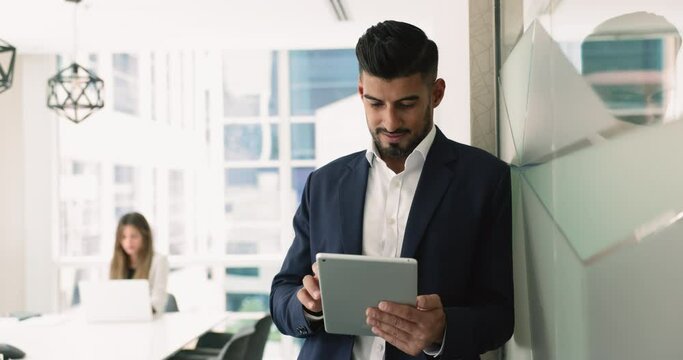 Smiling Indian businessman using tablet in office, reviewing editing documents, analyzing data using business analytics apps, managing schedules, make presentation for corporate clients or stakeholder