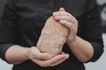 Unrecognizable woman in black coat with freshly baked delicious homemade ciabatta bread in hands in kitchen