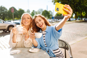 Beautiful woman friends using smartphone selfie together, while sitting at outdoor coffee shop. Fashion, beauty, blogging, tourism.