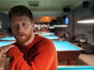 A young man holding a cue stick sits on a pool table watching in a pool bar at night