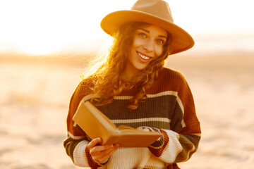 Young woman in a cozy sweater and hat enjoys nature, reading a book while sitting on a blanket at...