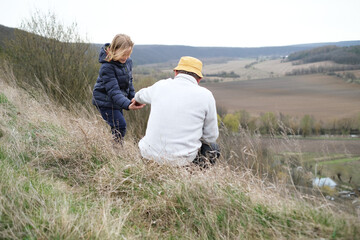Close up active senior and his grandson sitting on top of the hill and contemplating the landscape, turned back. Happy family active outdoor. Active activity in old age