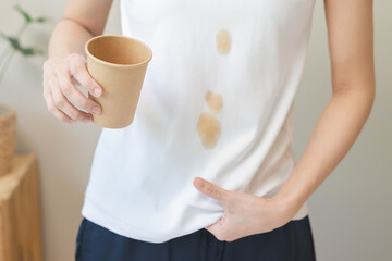 Cloth stain, disappointment asian young woman clumsy with hot coffee, tea stains on shirt, hand show making spill drop on white t-shirt, spot dirty or smudge on clothes at home, isolated on background - obrazy, fototapety, plakaty