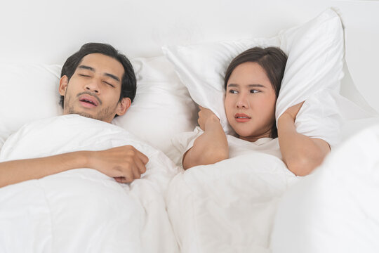 Snoring couple health asian man snore and sleeping at home while woman insomnia annoyed, bad noise cover ears with pillow, sleep problem apnea and relationship, wife looking at husband snoring in bed.